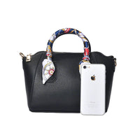 Front view of a black pebbled leather satchel handbag with a decorative nautical scarf-wrapped handle, shown with a smartphone to demonstrate compact size and capacity.