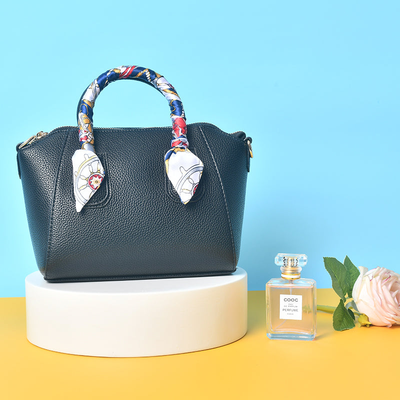 Black pebbled leather satchel handbag with scarf-wrapped handle, displayed on a pedestal with perfume and rose props for lifestyle product photography.